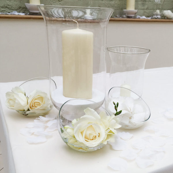 Hurricane lamp with large white candle inside, surroundd by three bowl vases with cream roses